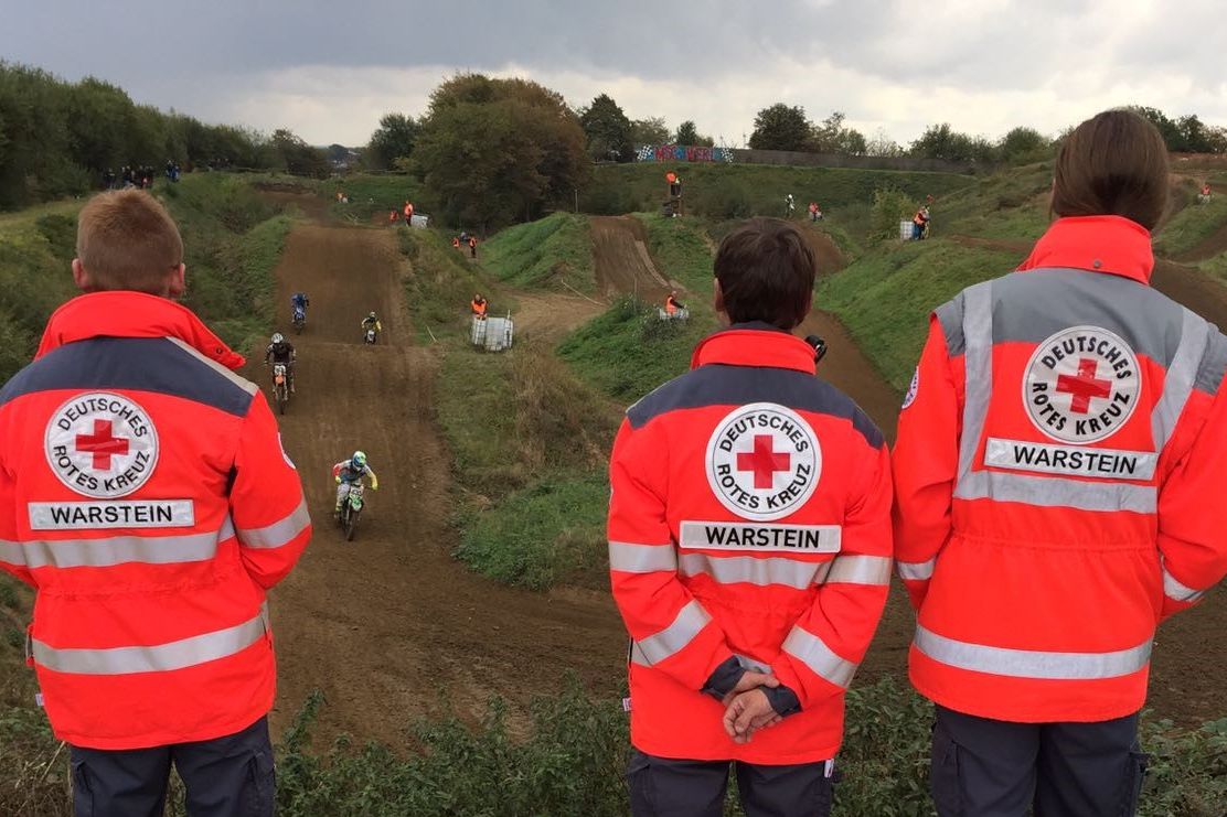 Drei Rotkreuzhelfer in Einsatzbekleidung stehen während eines Sanitätsdienstes auf einer Erhöhung und schauen auf eine Motorrad-Rennstrecke.